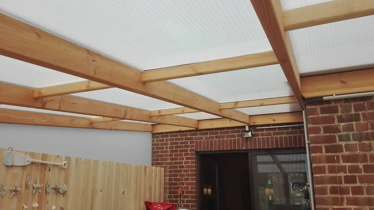 Charpente de terrasse en bois clair couverte de panneaux translucides, contre un mur en briques