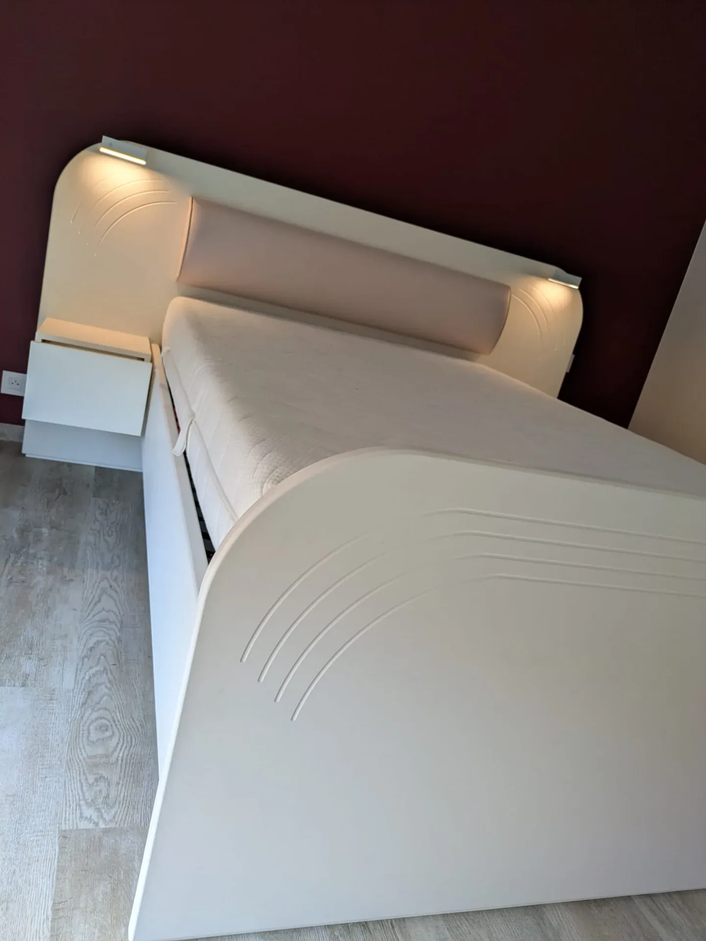 Lit sur mesure blanc cassé avec tête de lit arrondie, liseuses LED intégrées, traversin rose et chevet suspendu, devant un mur bordeaux