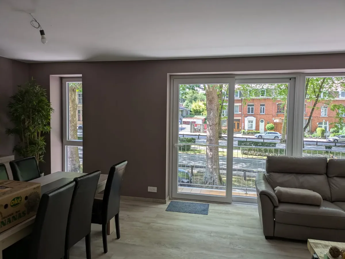 Salon avec une baie coulissante blanche ouvrant sur un balcon et une rue arborée, à côté d’une fenêtre haute et étroite