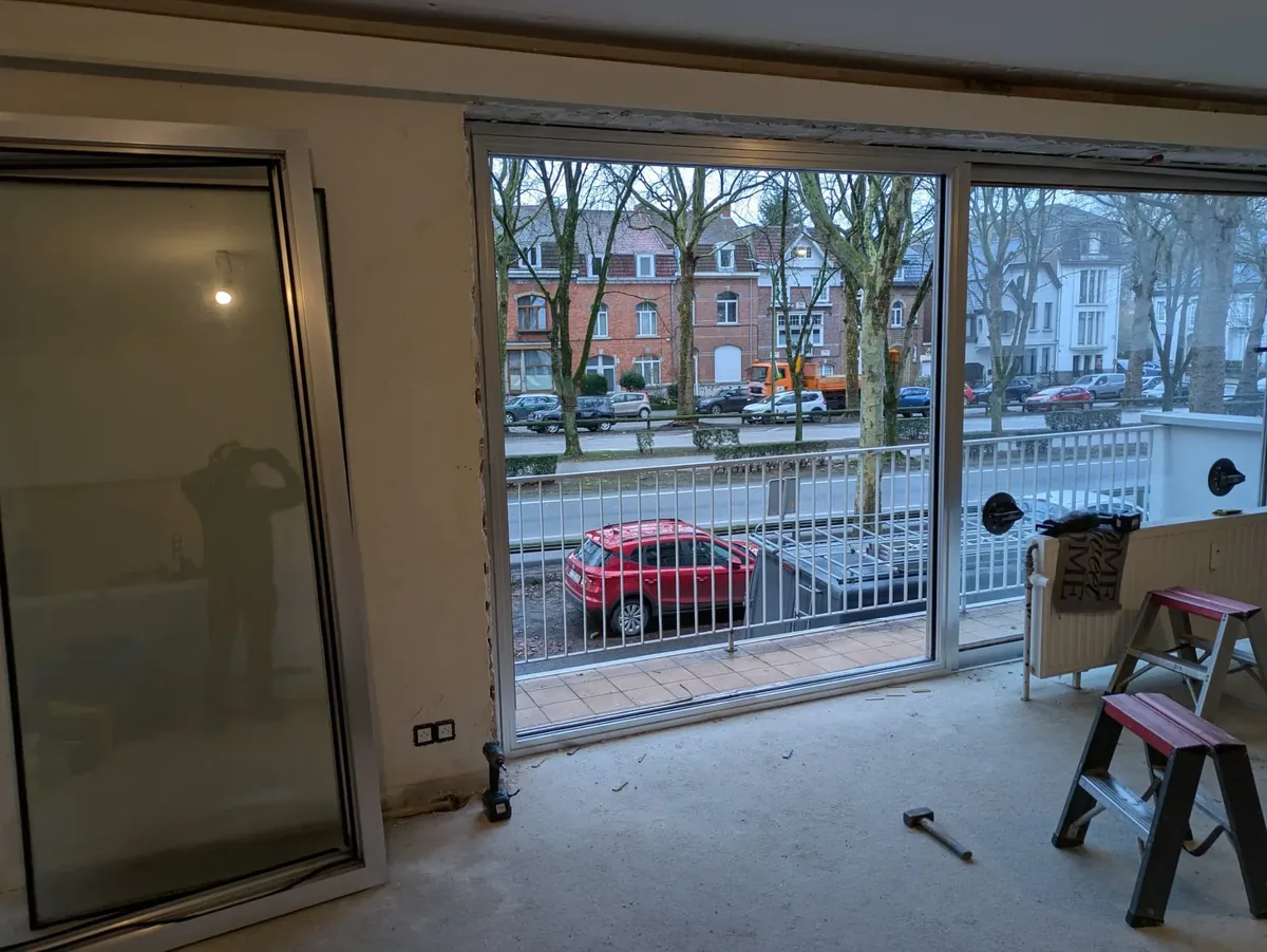 Salon en travaux : le cadre d’une grande baie coulissante posé dans l’ouverture, un vantail vitré appuyé contre le mur, échelles et outils au sol