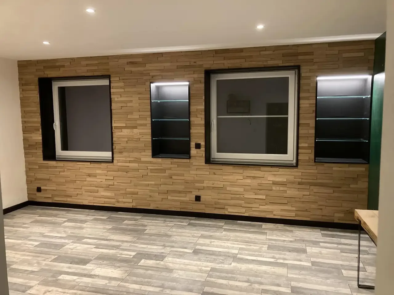 Mur habillé de lattes de chêne avec deux fenêtres et deux niches éclairées à encadrement noir, dans une pièce de vie au sol gris clair