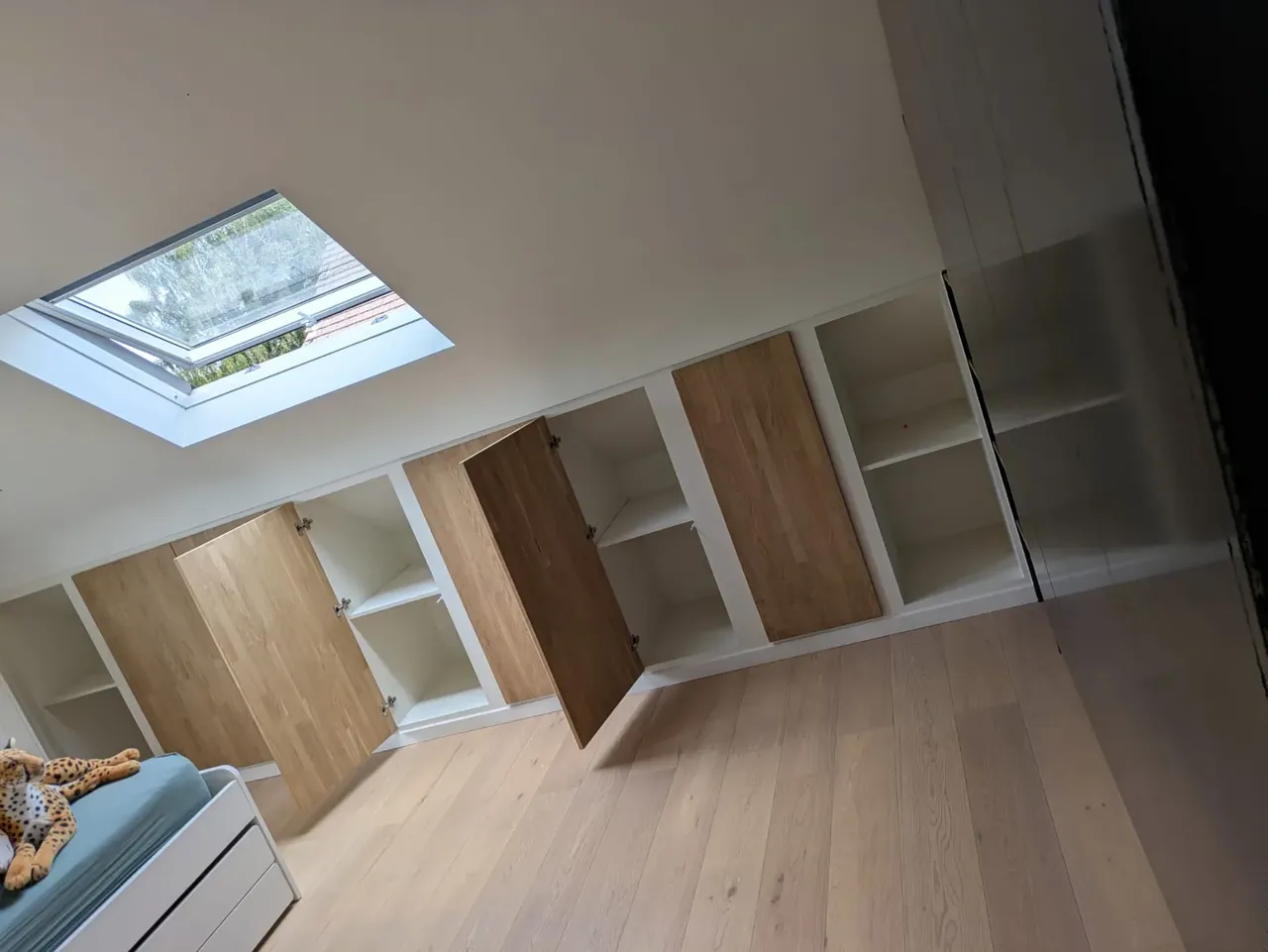 Rangements sous pente en blanc et chêne, portes ouvertes sur des étagères, avec une fenêtre de toit et un parquet clair