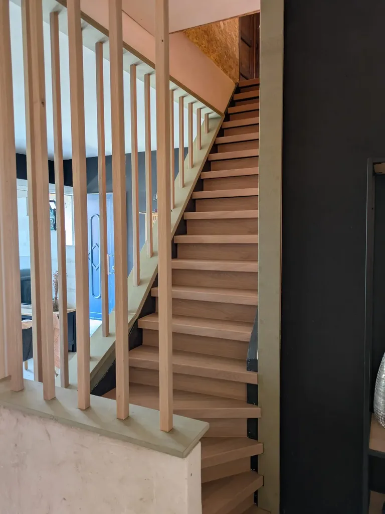 Escalier en chêne à marches tournantes, garde-corps à lames de bois verticales au premier plan, mur noir à droite