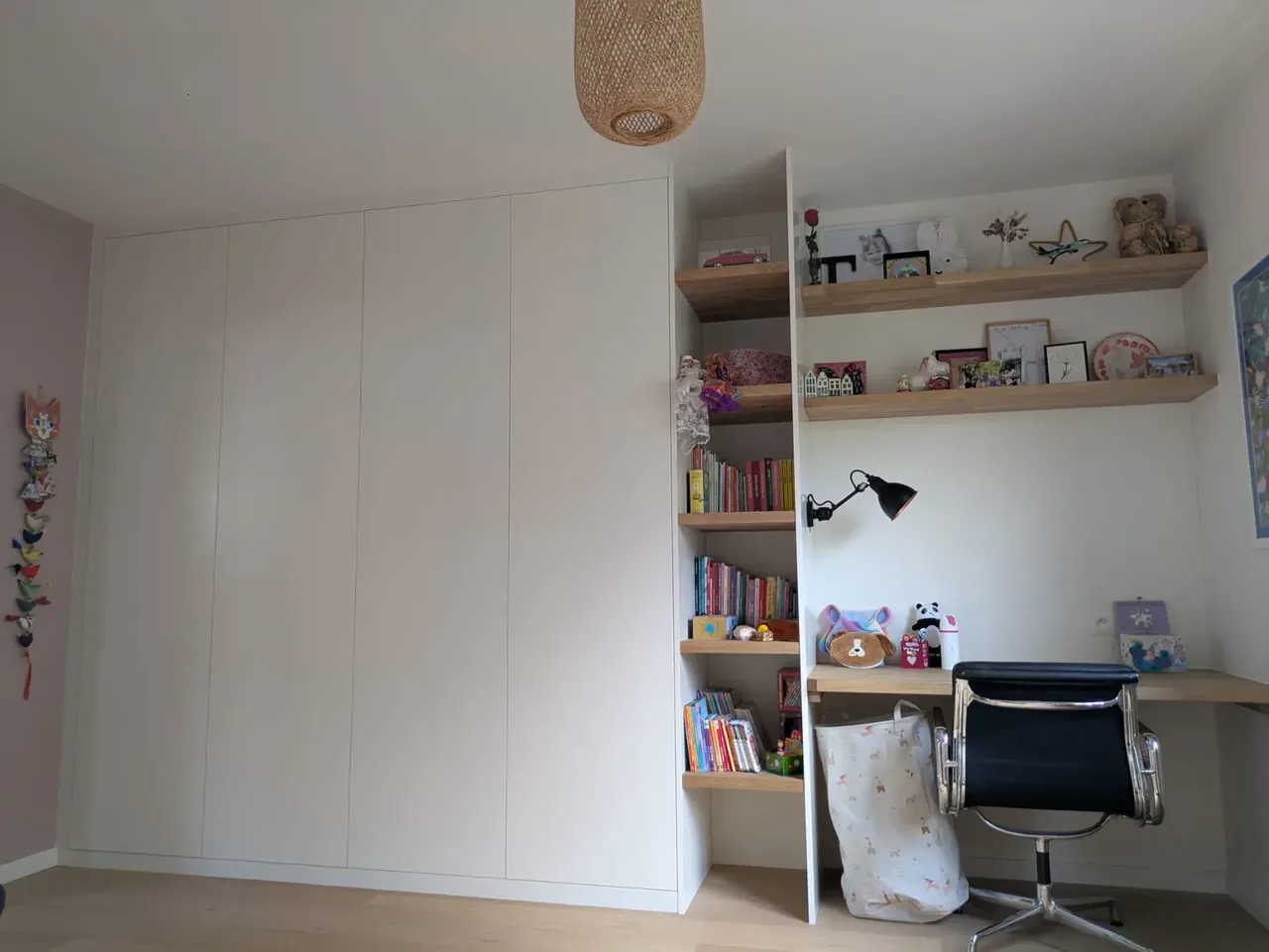 Chambre d’enfant avec armoire blanche à portes sans poignée, étagères et plan de bureau en chêne garnis de livres et de peluches, chaise de bureau