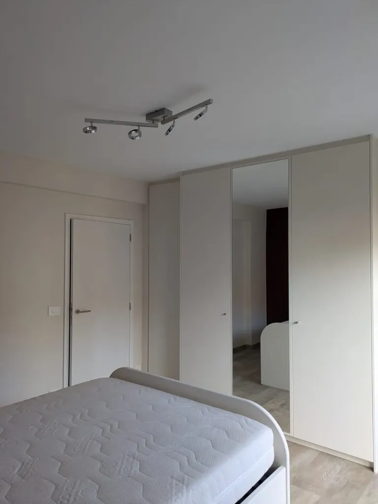 Chambre avec grande armoire-penderie blanc cassé à porte miroir, porte intérieure blanche et lit au premier plan