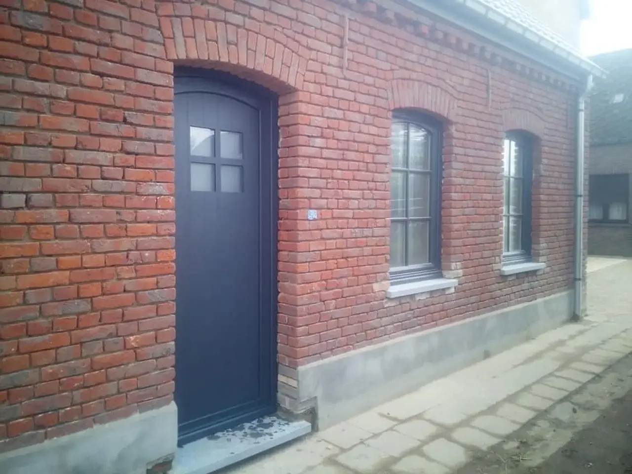 Façade en briques rouges avec une porte d’entrée cintrée anthracite à quatre petits carreaux et deux fenêtres à petits-bois