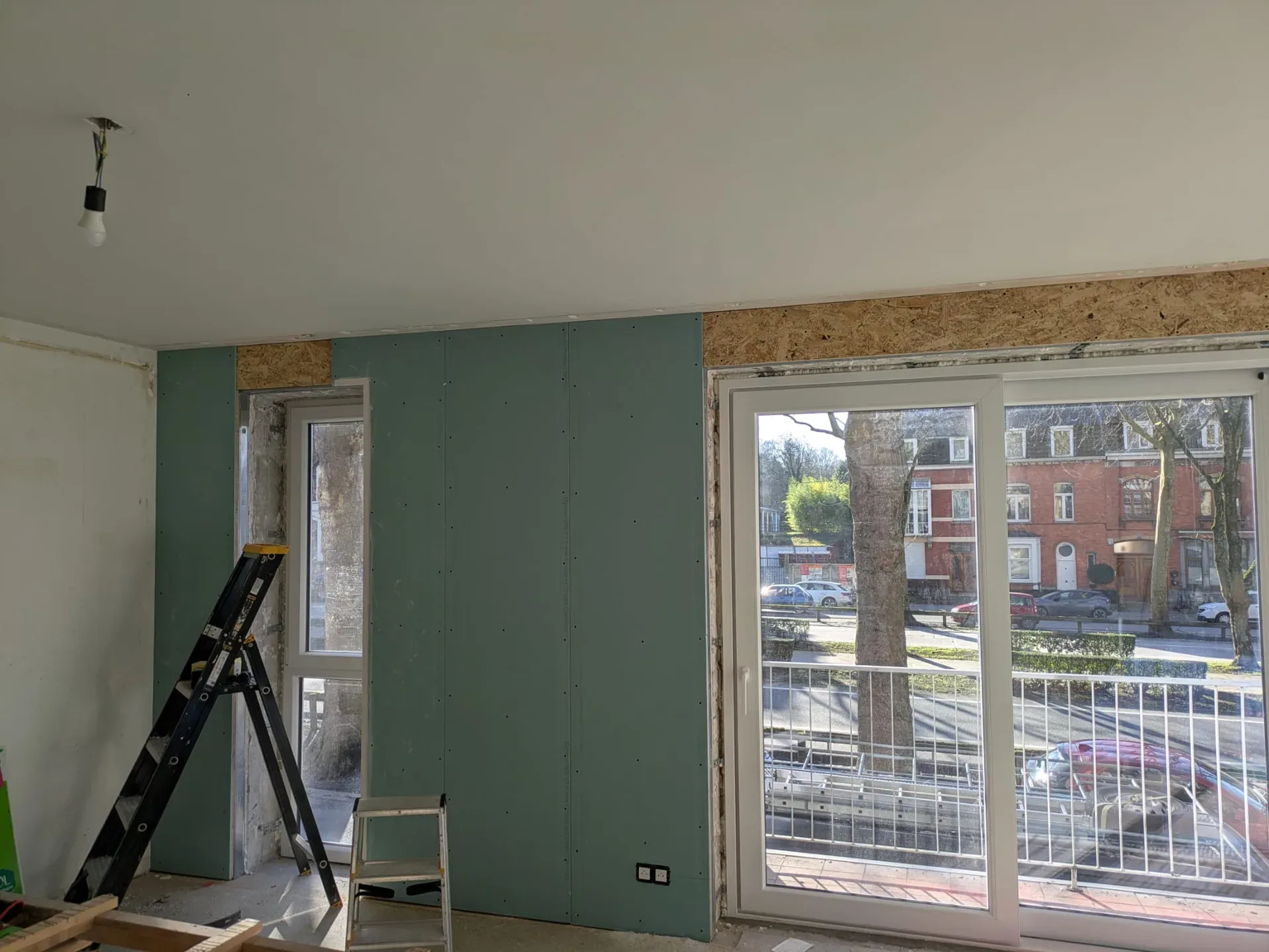 Pièce en travaux avec baie vitrée coulissante blanche installée, cloison en plaques de plâtre vertes et panneau OSB au-dessus de la baie