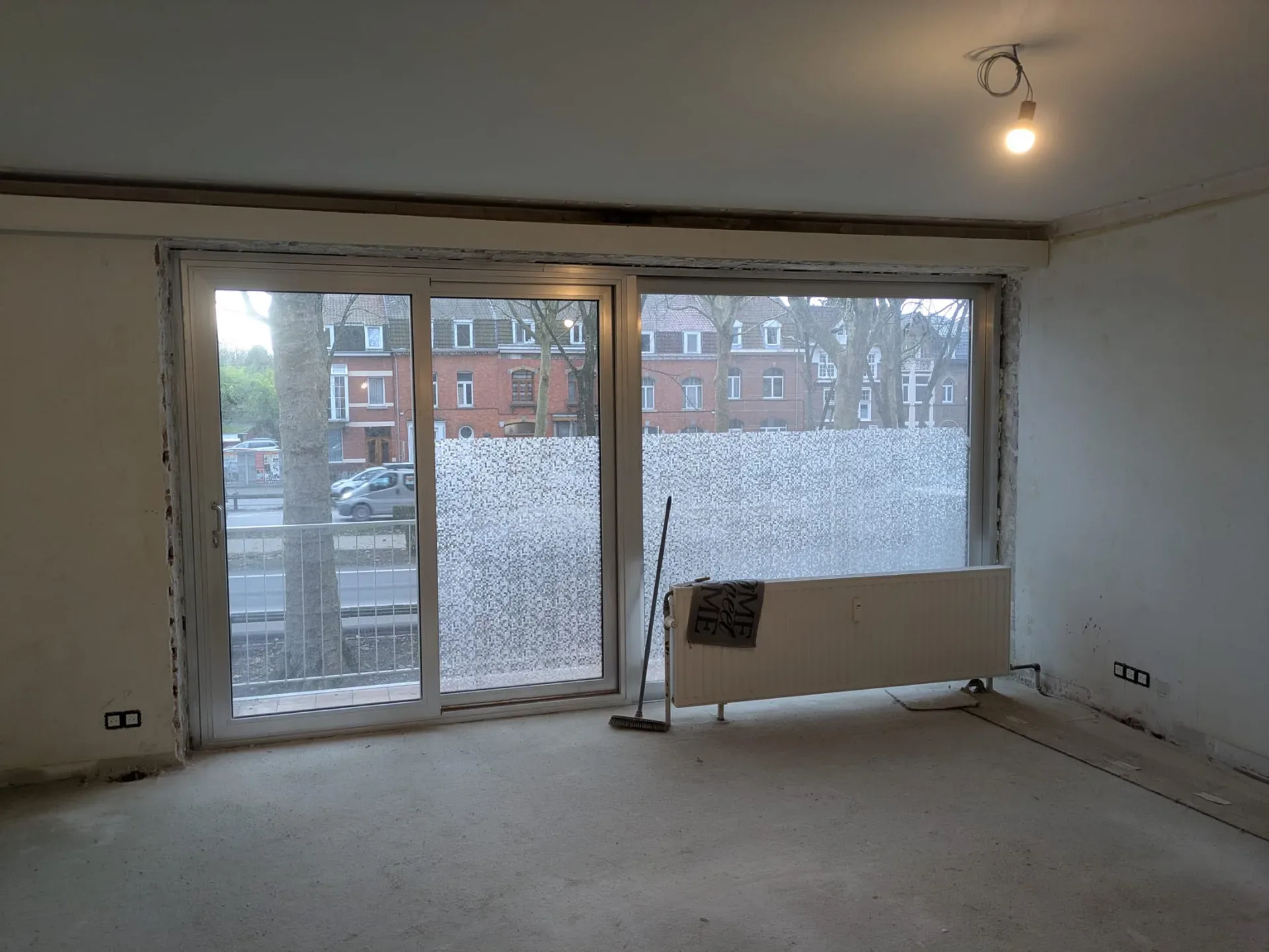 Pièce en travaux avec baie coulissante à deux vantaux installée, film dépoli sur la vitre, radiateur au sol, sol brut