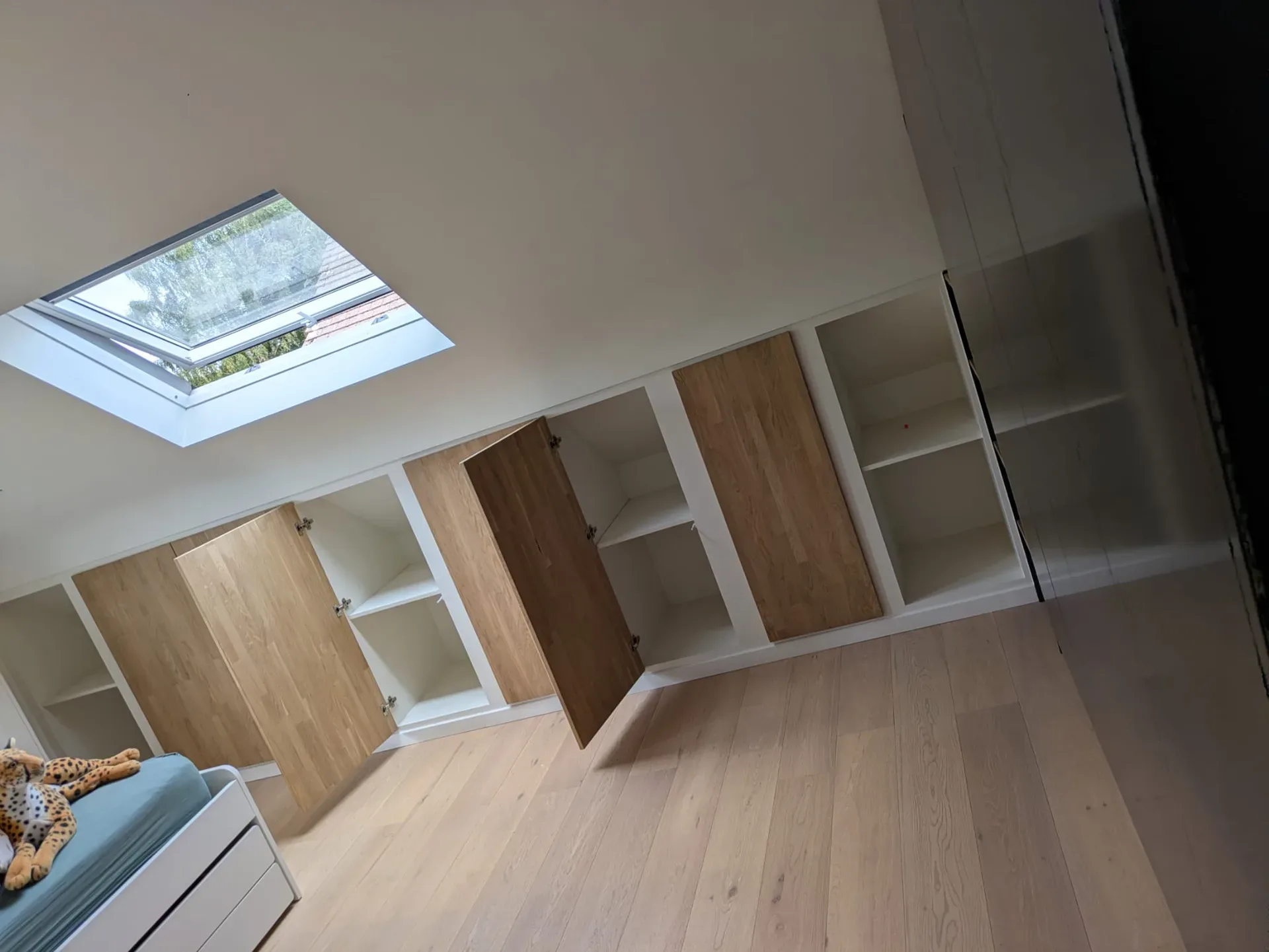 Rangements sous pente en blanc et chêne, portes ouvertes sur étagères, fenêtre de toit et parquet clair (cadrage penché)