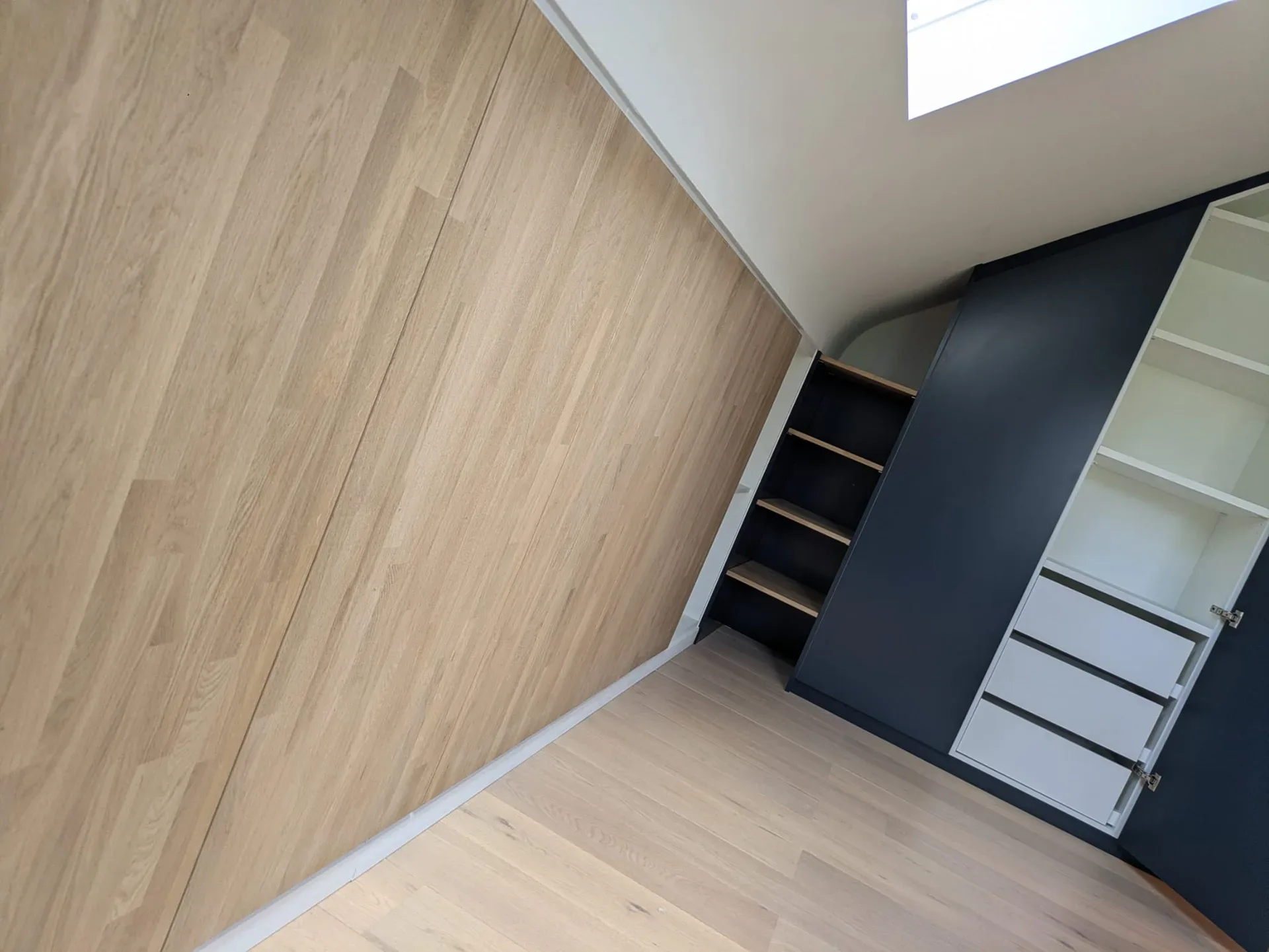 Façades de rangement en chêne sous pente, dressing bleu foncé avec tiroirs blancs et parquet chêne clair (cadrage penché)