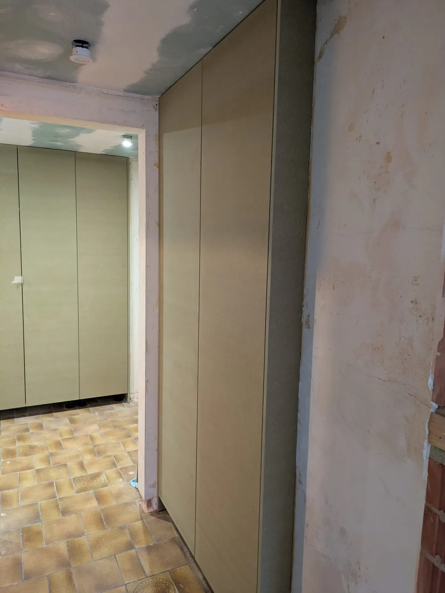 Façades d'armoires en panneaux bruts (non peints) dans un couloir et une pièce adjacente, plafond en cours de finition, sol carrelé
